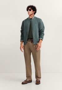 Grüne Bomberjacke mit weißen Streifen, olivfarbener Pullover, braune Hose und braune Loafers. Sonnenbrille vervollständigt den Look. Glatte Textur.