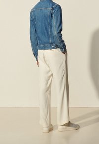 Denimová bunda v modré barvě s klasickým designem, doplněná bílými širokými kalhotami a světlými teniskami, z pohledu zezadu.