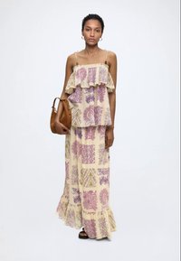 Femme portant une robe maxi crème avec des motifs floraux violets et verts, tenant un sac à main en cuir marron, debout devant un fond blanc.