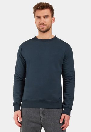 Man draagt een effen donkerblauwe sweater met ronde hals en grijze spijkerbroek, staand tegen een effen lichte achtergrond.