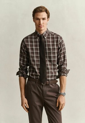 Homme portant une chemise à carreaux marron boutonnée avec les manches retroussées, une cravate noire, un pantalon marron, une ceinture et une montre-bracelet argentée, debout devant un fond uni.