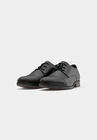 Zapatos de vestir para hombres en piel negra con cordones, diseño de puntera cap y tacón bajo apilado sobre un fondo blanco.