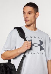 Grå t-skjorte med kort erm som har en svart logo og teksten "UNDER ARMOUR." En svart stropp fra en ryggsekk er synlig på skulderen.