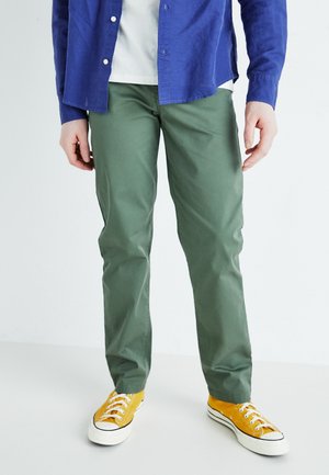 Homme portant un pantalon vert, des baskets en toile jaunes avec des semelles blanches, une chemise blanche et une chemise bleue boutonnée, debout sur un sol blanc.