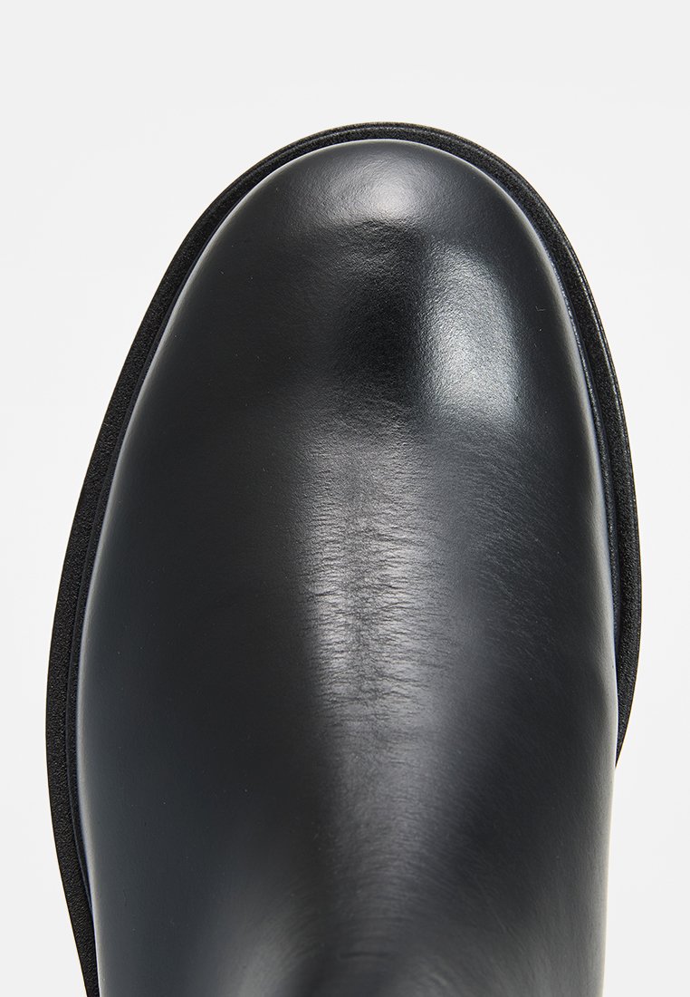 Bottine en cuir noir avec une finition lisse et brillante. Dotée d'un bout arrondi et de coutures minimales. La semelle a un léger bord surélevé.
