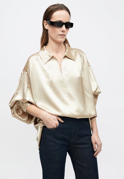 Bluza din satin, auriu deschis, cu guler, umeri căzuți și mâneci voluminoase. Asortată cu blugi din denim închis și ochelari de soare negri, dreptunghiulari.