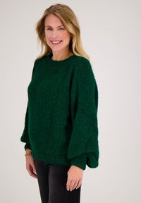 Vrouw met blond haar die lacht en gouden oorringen draagt, een dikke donkerblauwe trui, zwarte broek en gouden armbanden, op een witte achtergrond.