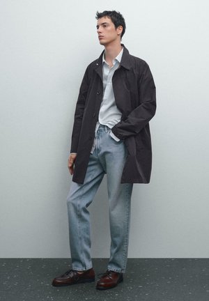 Manteau léger marron foncé sur une chemise boutonnée bleu clair, associé à un jean en denim clair et des chaussures foncées. Texture lisse et coupe ajustée.