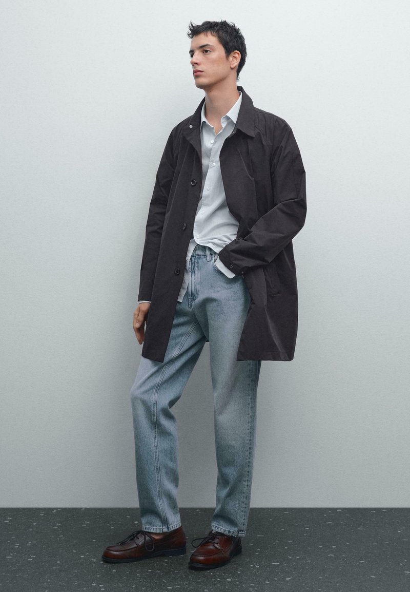 Cappotto leggero marrone scuro indossato sopra una camicia a bottoni azzurro chiaro, abbinato a jeans in denim chiaro e scarpe scure. Tessuto liscio e taglio su misura.