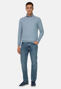 Homme portant un pull bleu clair par-dessus une chemise à col, un jean de teinte moyenne et des baskets bleu marine, debout avec une main dans la poche.