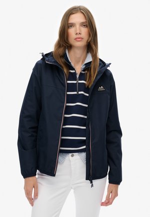 Veste coupe-vent marine avec une fermeture éclair à l'avant et des poignets élastiques. Présente un petit logo sur la poitrine et est portée sur un pull rayé.