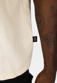 Beige cotton t-shirt with a straight hem and a black tag at the side featuring a white geometric logo. Exposed arm with tattoos visible.