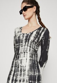 Black and white long-sleeve dress with a dotted pattern, scoop neckline, and fitted design. Model wears black rectangular sunglasses.