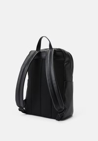 Sac à dos noir avec un dos en maille texturée, des bretelles rembourrées, des côtés en cuir lisse et une fermeture supérieure zippée. Poche latérale visible.