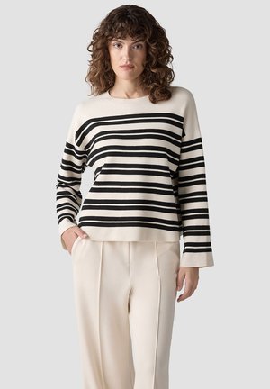 Frau mit lockigem Haar trägt einen cremefarbenen Pullover mit schwarzen horizontalen Streifen und cremefarbene Hosen mit weitem Bein, steht mit einer Hand in der Tasche.