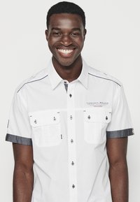 Camisa blanca de manga corta con cuello tipo botón, dos bolsillos frontales, detalles en color plata y acentos en azul oscuro en las mangas y los hombros.