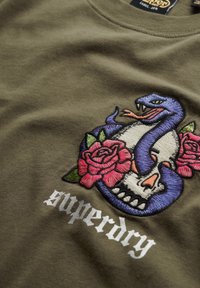 Olivengrøn bomulds t-shirt med et broderet design af en lilla slange og kranie, med pink roser og hvid "superdry" tekst.