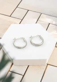 Silverörhängen i form av hoops med ett geometriskt mönster längs bandet, utrustade med en gångjärnsknapp för säker användning, visade på en vit hexagonal bas.