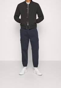 Veste bomber noire avec des boutons-pression à l'avant, pantalon cargo bleu marine avec poches latérales et baskets blanches avec des accents gris. Design simple et décontracté.
