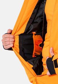 Giacca arancione brillante con fodera nera, caratterizzata da un'apertura con zip e una tasca interna progettata per la conservazione. Tessuto strutturato visibile.