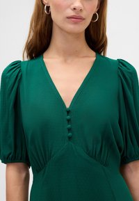 Roheline kleit sügava V-kaelusega, puffy varrukatega ja dekoratiivsete nööpidega. Tekstuurse kangaga, millel on kerge läige, ning kogutud vöödetail.