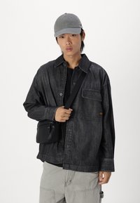 Camicia di denim nera con taschino, indossata sopra una maglietta nera. Cappellino grigio e piccola borsa a tracolla nera. Pantaloni grigi chiari, vestibilità rilassata.