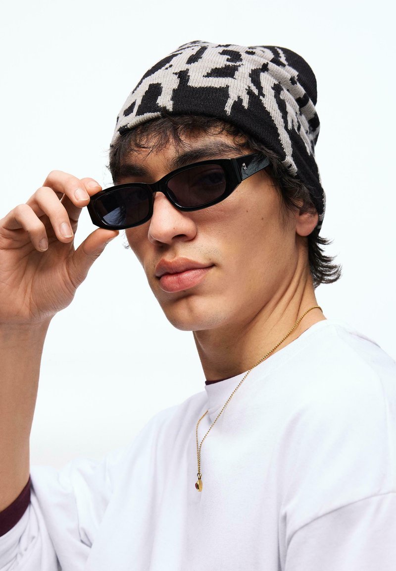 Jeune homme ajustant des lunettes de soleil rectangulaires noires, portant une chemise blanche, un bonnet noir et blanc à motifs, et un collier en or avec un pendentif.