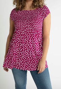 Mujer vestida con una blusa corta de manga con flores en magenta y un top fruncido, con jeans azules, de pie contra un fondo liso.