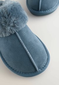 Blå ruskindstøfler med blød pelskant, med en syet søm og rundet tådesign, hvilende på en lys baggrund.