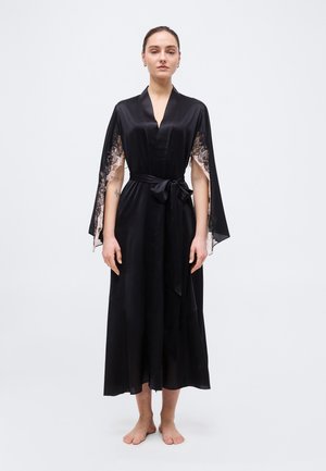 Γυναίκα όρθια, ξυπόλητη, φορώντας μαύρη σατέν ρόμπα με μανίκια-κάπα διακοσμημένα με δαντέλα και ζώνη στη μέση, μπροστά σε απλή φόντο.