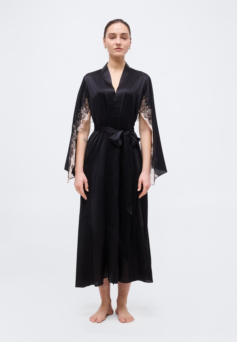 Γυναίκα όρθια, ξυπόλητη, φορώντας μαύρη σατέν ρόμπα με μανίκια-κάπα διακοσμημένα με δαντέλα και ζώνη στη μέση, μπροστά σε απλή φόντο.