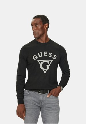 Suéter negro de punto con un destacado logo gris "GUESS" y un diseño de triángulo en la parte frontal. Combinado con pantalones de mezclilla grises.