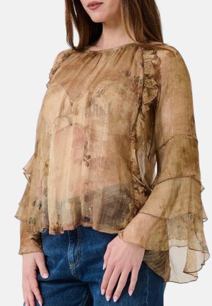 Donna che indossa una camicetta marrone trasparente a maniche lunghe con volant e righe metalliche, abbinata a jeans blu, mostrata dalle spalle ai fianchi.