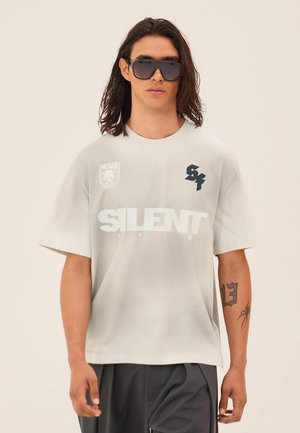Mężczyzna z długimi włosami, noszący duże okulary przeciwsłoneczne, jasnoszary t-shirt z nadrukiem „SILENT” oraz ciemne spodnie, stojący na tle jednolitego tła.
