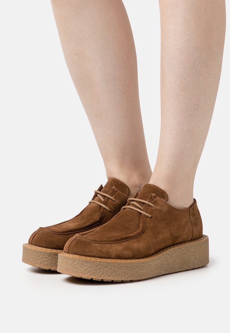 Chaussures à plateforme en daim marron à lacets portées sur des pieds à peau claire sur fond blanc.