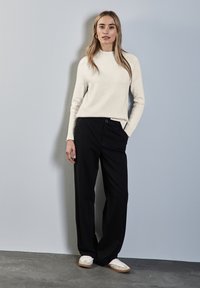 Crème trui met een geribbelde halslijn, gecombineerd met zwarte wijde broek. Model draagt witte sneakers met beige accenten. Eenvoudige achtergrond.