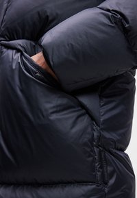 Casaco puffer em azul-marinho escuro com uma textura suave e brilhante. Apresenta uma manga grande e almofadada e um bolso lateral com fecho de pressão.