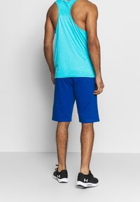 Maschio con top atletico senza maniche blu, pantaloncini blu reale e scarpe da corsa nere Under Armour, visto da dietro su sfondo bianco.