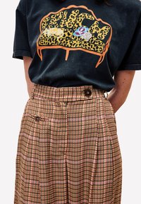 Zwart T-shirt met kleurrijk luipaardpatroon en grafische tekst; high-waisted geplooide bruine geblokte broek met knoopdetail.