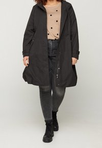 Cappotto impermeabile nero con cappuccio, con zip frontale, tasche laterali e vestibilità comoda, indossato sopra una maglietta beige con pois neri.