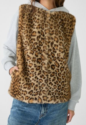 Persoon draagt een nep bont vest met luipaardprint over een lichtgrijze capuchontrui en blauwe spijkerbroek, met één hand in het vestzakje.