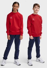 Felpe rosse a maniche lunghe con collo rotondo indossate da una ragazza e un ragazzo, abbinate a pantaloni blu navy e scarpe da ginnastica bianche. Logo visibile sulle felpe.