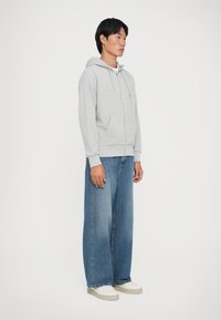 Sudadera gris con capucha y bolsillos tipo canguro en la parte frontal, combinada con jeans de mezclilla azul de pierna ancha y zapatillas blancas, de pie contra un fondo liso.