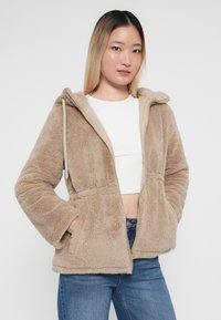 Veste à capuche zippée beige duveteuse avec une texture douce, deux poches avant et une doublure beige clair contrastante. Portée par-dessus un haut blanc et un jean.