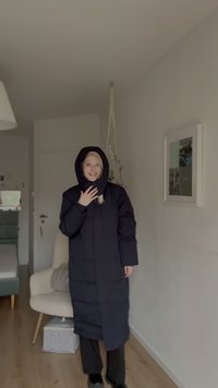 Ilgas juodas pūkinis paltas su gobtuvu, su siuvinėta tekstūra, šoninėmis kišenėmis ir iki kelių ilgio dizainu. Dėvimas ant tamsių kelnių paprastoje vidaus aplinkoje.