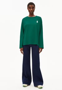Groen T-shirt met lange mouwen en een wit lintlogo, gecombineerd met wijde donkerblauwe jeans en witte sneakers met groene accenten.