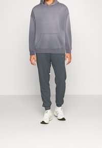 Henkilö, jolla on harmaa huppari ja tummanharmaat collegehousut sekä valkoiset lenkkarit, seisomassa yksinkertaisen vaalean taustan edessä.