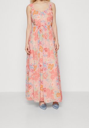 Femme portant une robe longue rose sans manches avec un imprimé floral multicolore et des chaussures pointues bleues avec décoration florale.