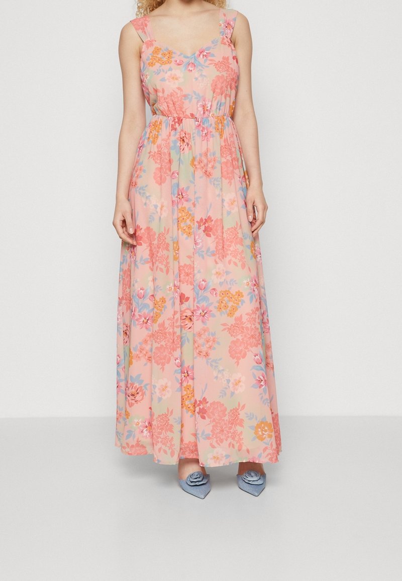 Femme portant une robe longue rose sans manches avec un imprimé floral multicolore et des chaussures pointues bleues avec décoration florale.