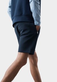 Pantaloni corti blu navy in un materiale morbido con tasche laterali. Presentano una chiusura con zip sulla tasca destra e una texture liscia.